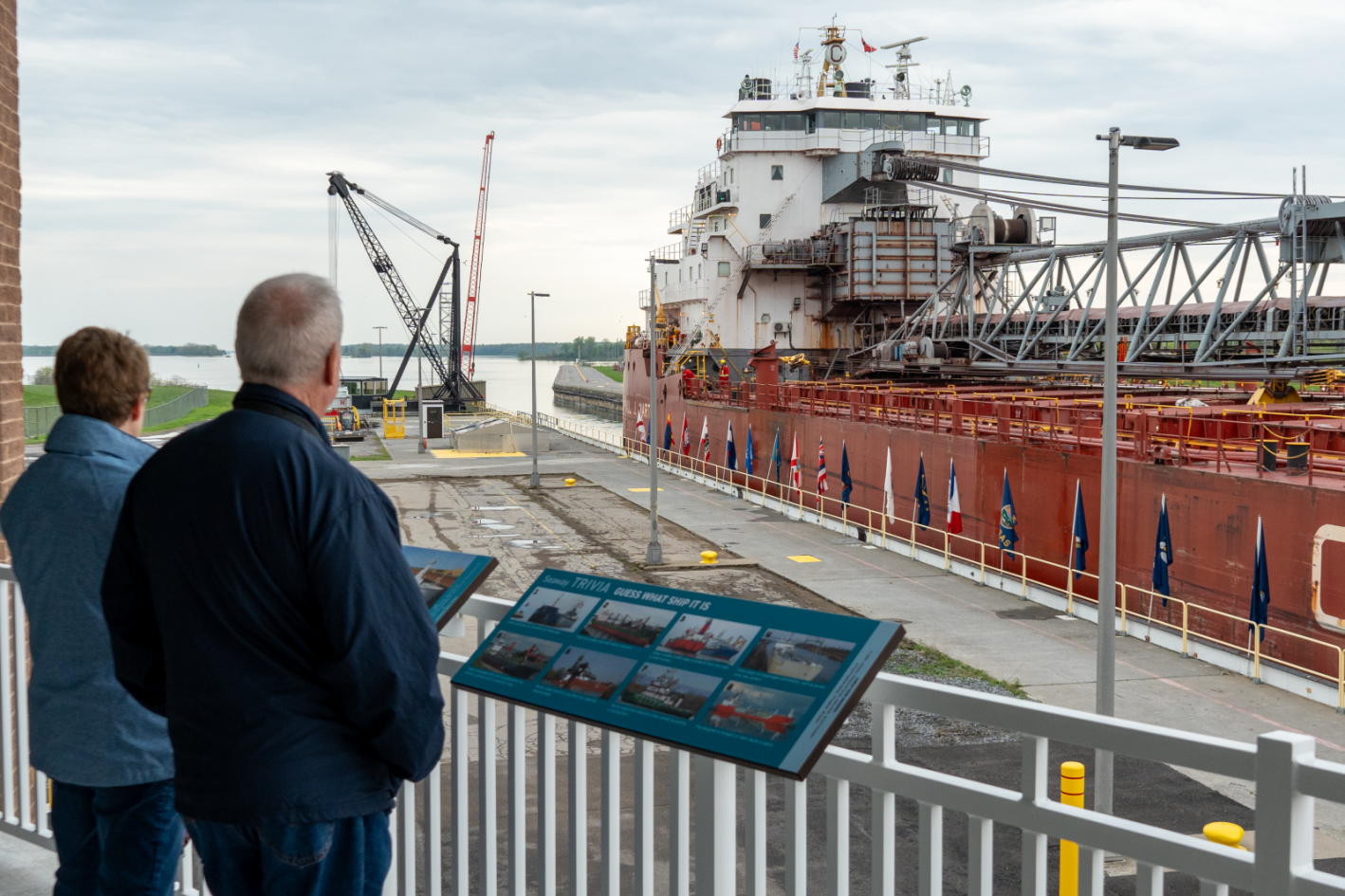 Two visitors on an elevated observation deck at the Seaway Visitor Center looking at a large red freighter in Eisenhower Lock. Interactive trivia signs are mounted to the railing in the foreground.